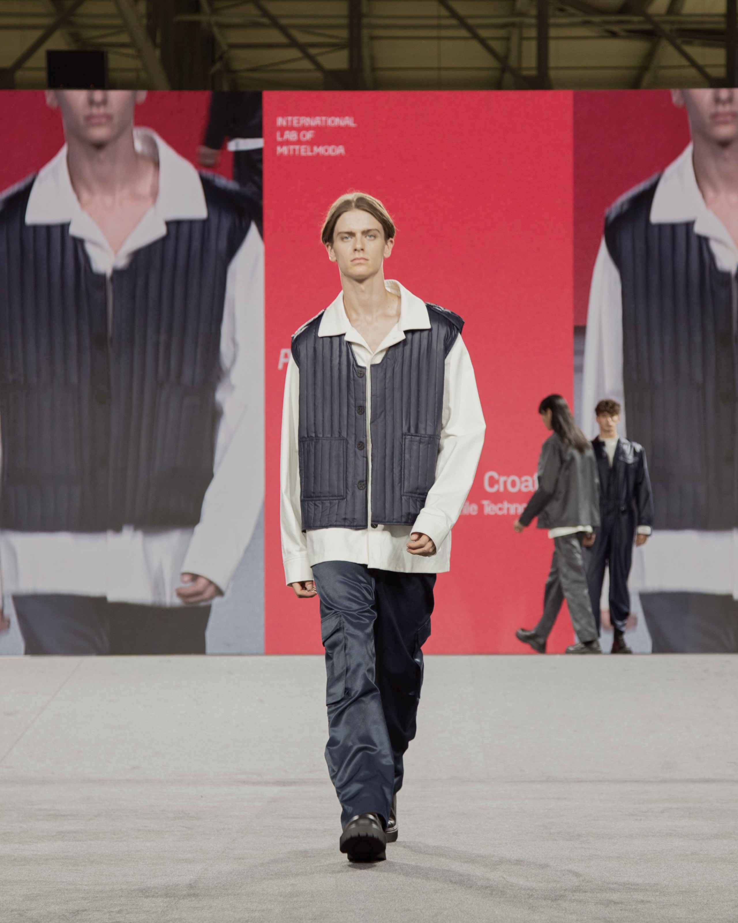 Model wearing blue vest, pants, and white shirt from KOVARI collection on Mittelmoda show.
